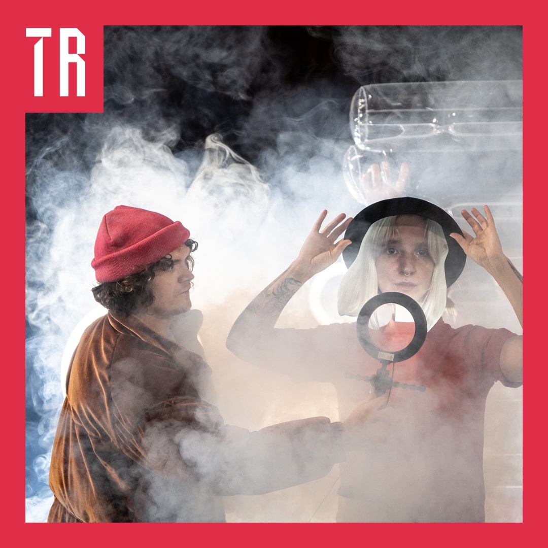 Two figures in a smokey room. One holds a ring light in front of the face of another. They are both wearing hats.