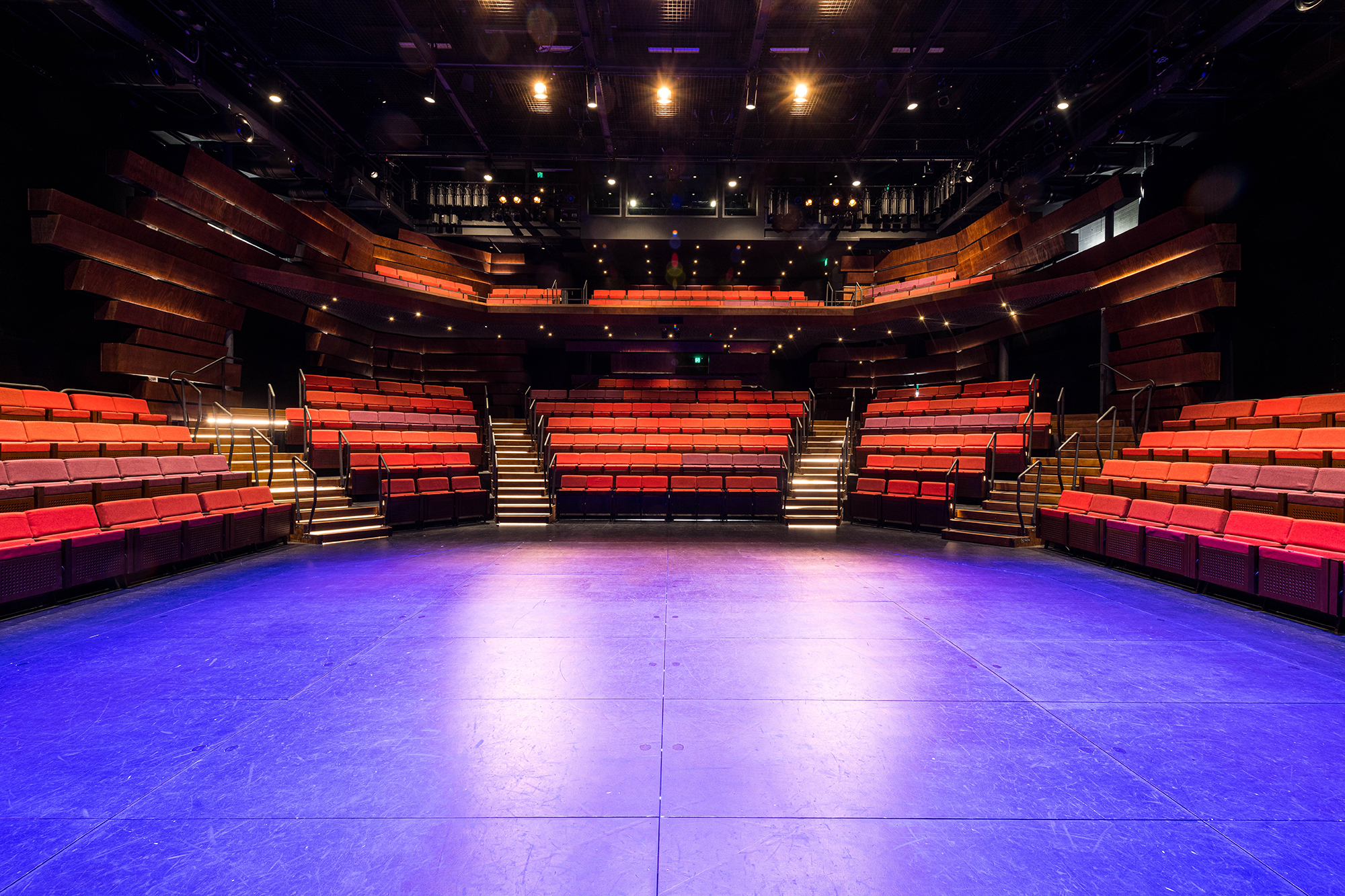 A photo of The Theatre Royal's Studio Theatre at The Hedberg. A blue light hits the stage and the point of view is of the performer on stage looking into the red seating banks.