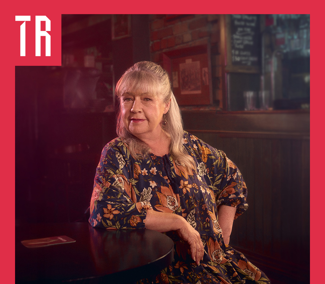 Photo: Mia McDonald A woman sits on a chair, leaning her arm on a dark woodern table. She is smiling, in a wise way. She wears a floral dress of black and tan colours. Her hair is greying blonde. She look as if she is about to tell us a story
