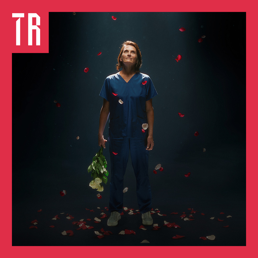 Image: Mitch Osborne, Oi Studios A woman stands against a dark background, she is wering blue hospital scrubs and is holding a bunch of flowers. Petals fall from above her.