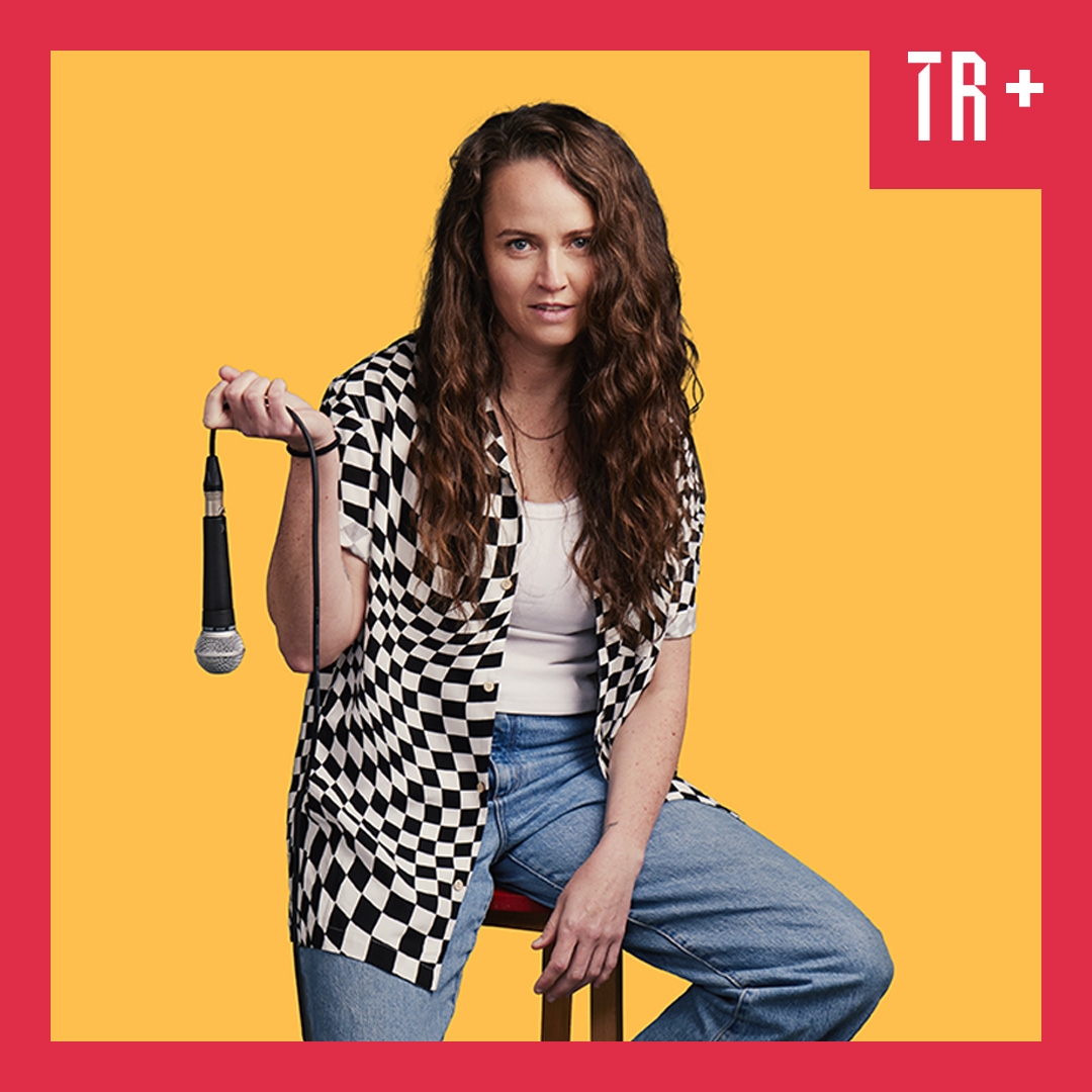 Image: supplied A portrait of comedian Zoe Coombs Marr, sitting on a wooden stool, slouching, wearing blue demin jeans and white tshits with a black and white checked shirt open at the fron. They hold a mic and have long wavy hair