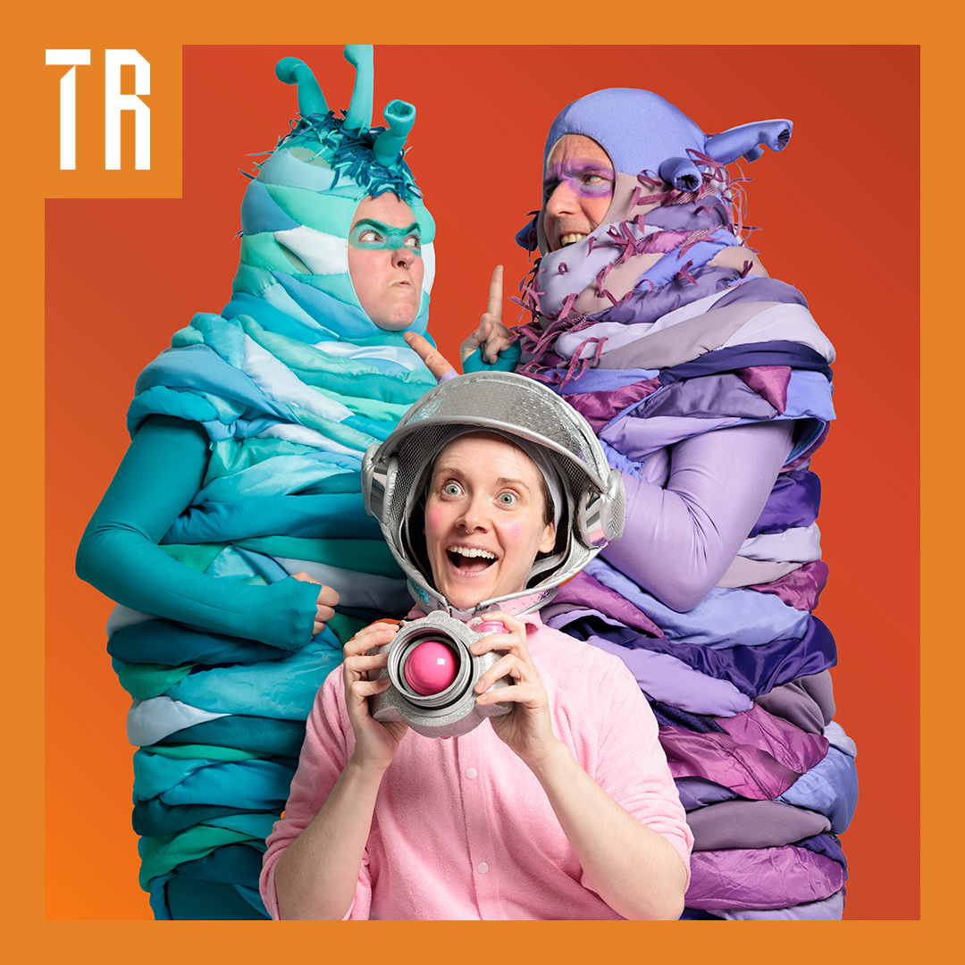Image: supplied A colourful photo of three performers dressed in costumes, two a dressed as worm like aliens and one like and explorer.