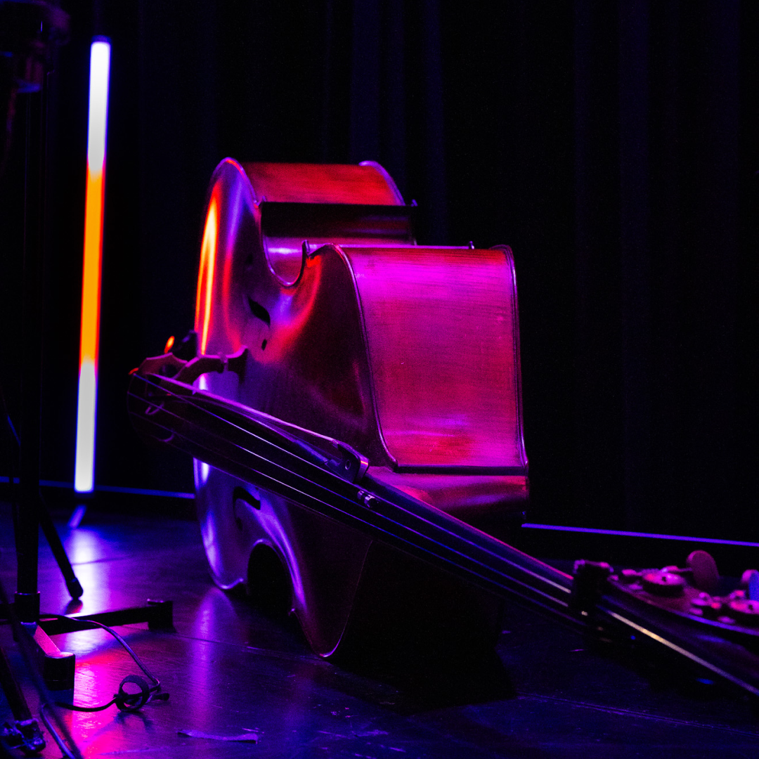 Image: supplied A cello illuminated by vivid purple and red stage lighting, resting on a dark stage beside a vertical light bar.