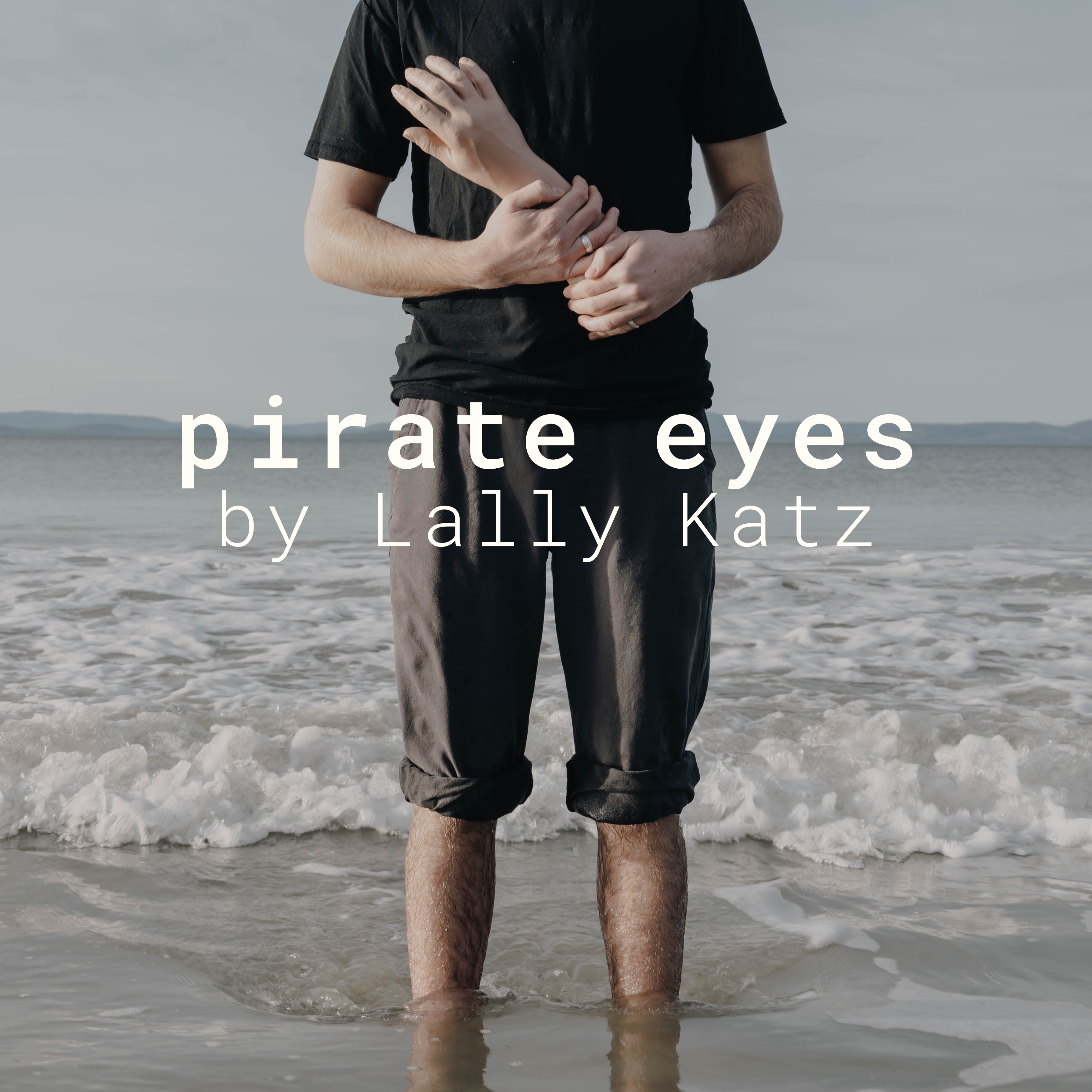 Pirate Eyes by Lally Katz A person in a black tshirt and black pants. Their pants are rolled up and they are standing in ankle deep ocean water. A small wave is crashing behind them. They are holding a fake hand.