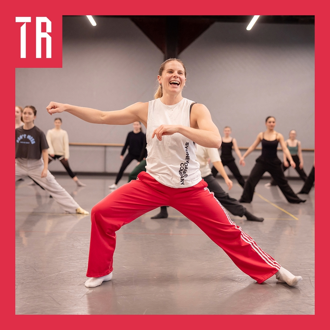 Photo: Daniel Boud A woman wearing exercise clothing raises her right arm out. Her legs are far apart and her right leg is bend. She is laughing and there are multiple people behind her doing the same thing. They look to be in a rehearsal room.