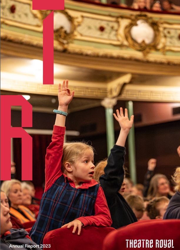 A girl sits in a crowded theatre, her hand raised in the air like she wants to ask a question